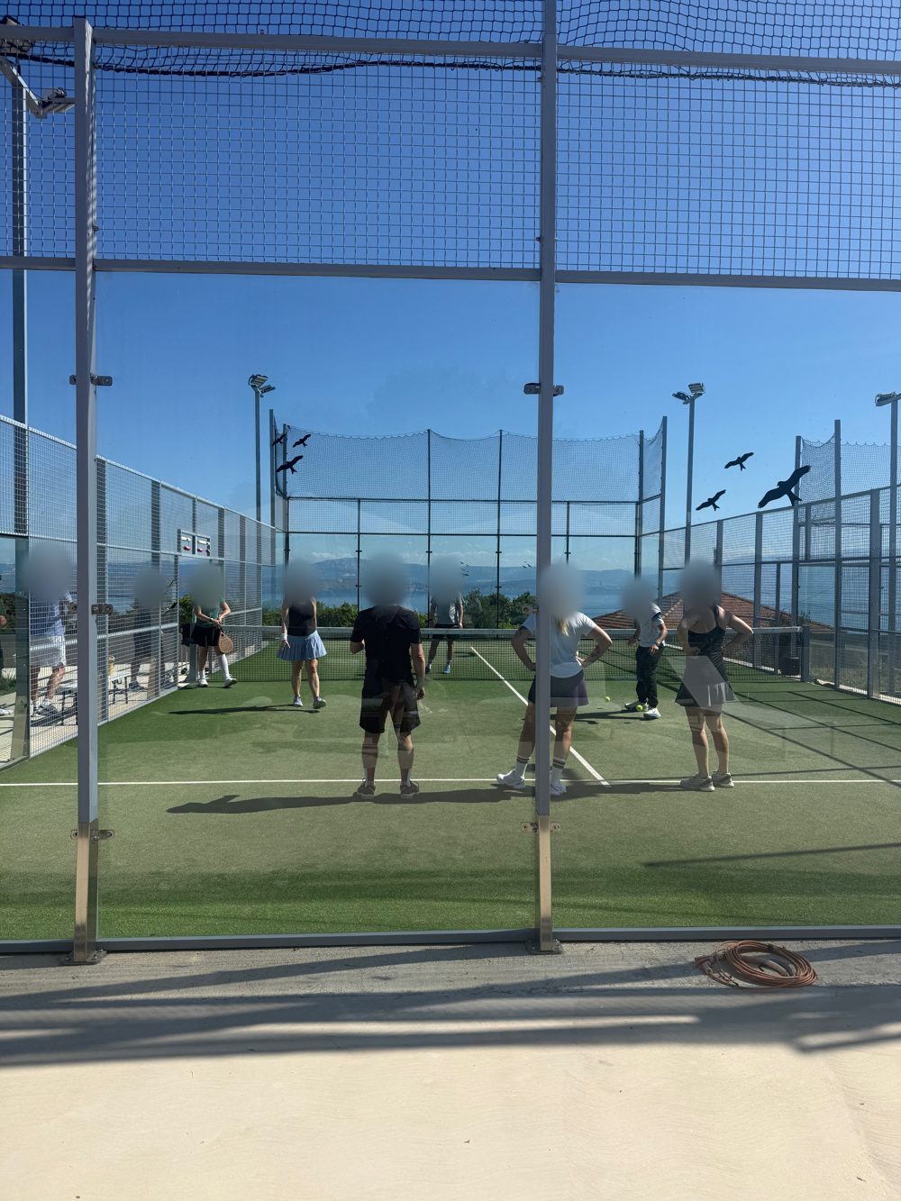 Padelbane med net og havet bag hegnet, spillerne står med ansigterne sløret