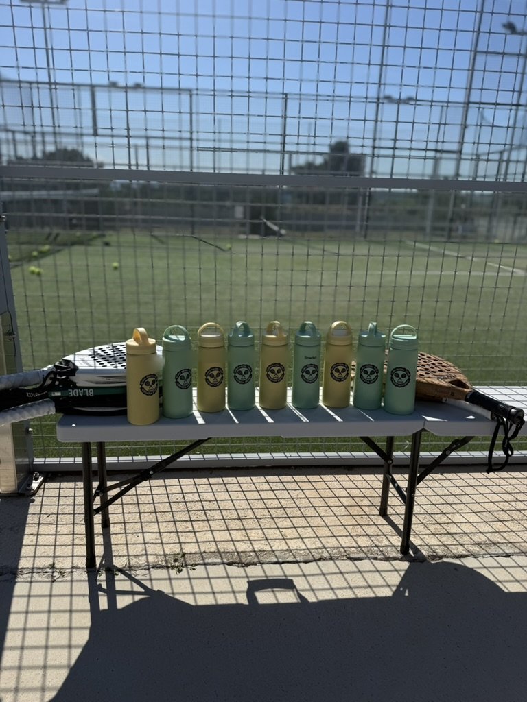Ni drikkedunke med logoet står på række på et bord ved padelbanen