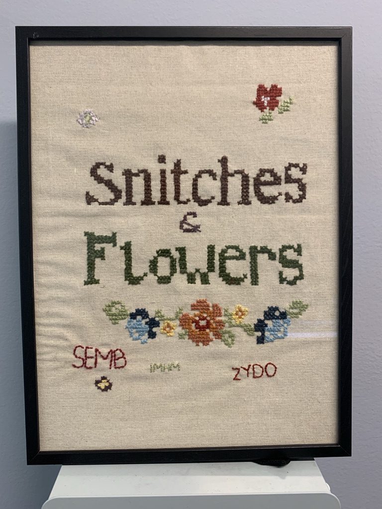 Indrammet broderi med teksten Snitches & Flowers, blomster og tre sæt initialer